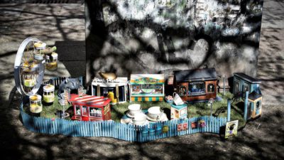 Ett tivoli i miniatyrformat. Tivolit har flera små bodar och ett pariserhjul. Det är omgärdat av ett blått staket och över ingången är en skylt där det står ”Tjoffsans tivoli”.