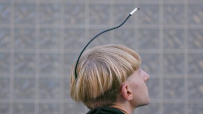 Ett huvud bakifrån på en kille. Han har blont tjockt hår. Ur hans bakhuvud sticker det ut en svart pinne som sträcker sig fram över hans huvud och slutar strax framför hans panna.