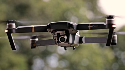 En drönare flyger i luften. Den är grå med fyra pinnar som sticker ut åt varsitt håll. Längst fram syns en kameralins.