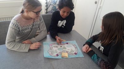 Tre elever sitter runt ett bord i ett klassrum och spelar ett spel. Spelet föreställer en stig runt några träd.