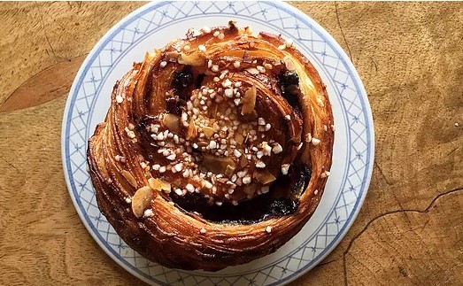 En bulle med pärlsocker och mandel på. Bullen ligger på ett fat med blå kanter. Fatet står på ett yta av grovt trä.