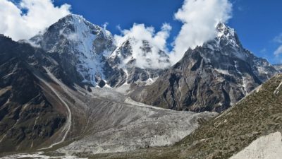 Några berg syns mot en blå himmel. Bergen är väldigt höga och har snö högst upp. Runt topparna ligger det vita moln.