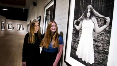 Två personer framför ett foto i ett galleri. I galleriet hänger fotografier. Ett fotografi är på en av personerna som står i galleriet. Personen på fotografiet har en ljus klänning. Personen håller i sitt hår.