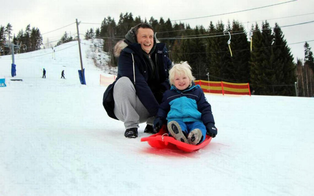 Ett barn sitter på en röd pulka i snötäckt backe. Bakom barnet sitter en vuxen på huk och skrattar.