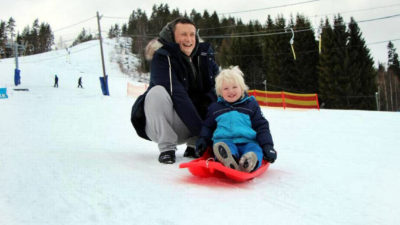 Ett barn sitter på en röd pulka i snötäckt backe. Bakom barnet sitter en vuxen på huk och skrattar.