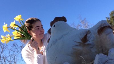 Ett barn håller i en bukett med påskliljor. I förgrunden en höna i porslin. Klarblå himmel.