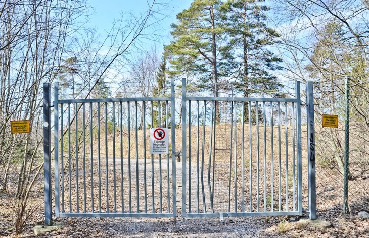 En grind och ett staket med skyltar som säger att det är förbjudet att gå in. Innanför grinden står träd vid sidan om en grusväg.