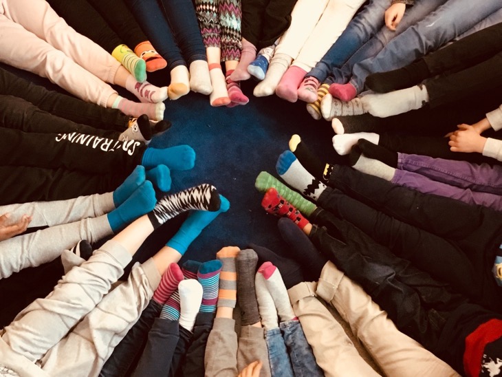 Barn sitter i ring med fötterna tillsammans. Strumporna har olika mönster och färger. Mattan under dem är mörkt blå.