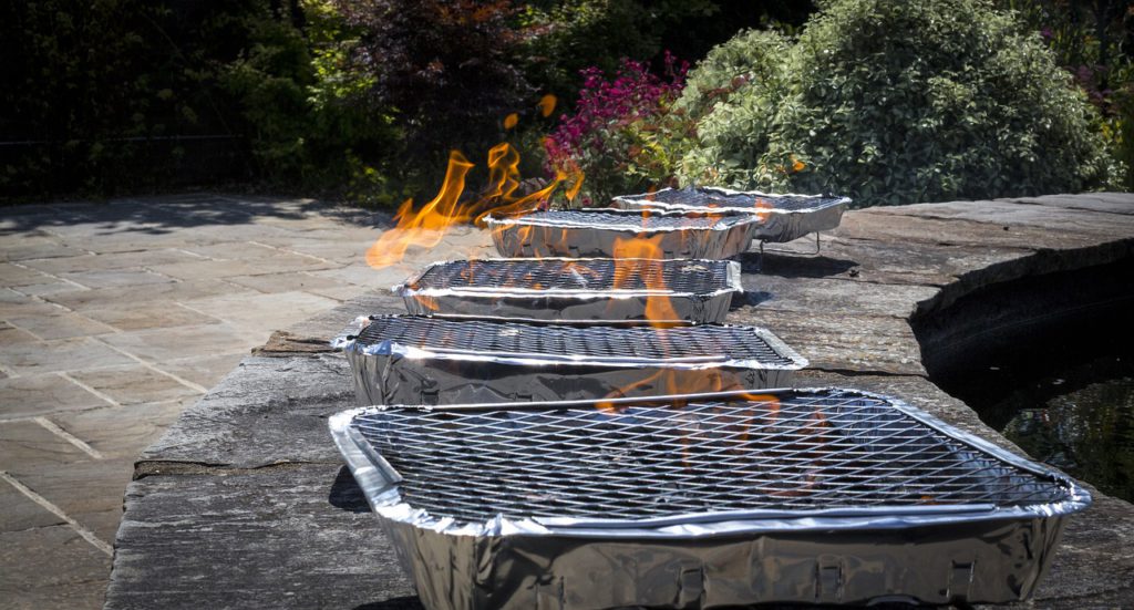 Bild på fem engångsgrillar som står uppradade på en stenmur. Det kommer lågor från grillarna.