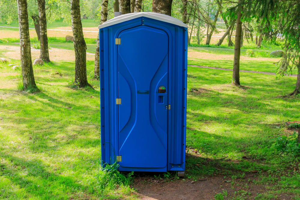 En bajamaja står på en gräsmatta. Bakom syns flera träd. Bajamajan ser ut som en hytt av plast.